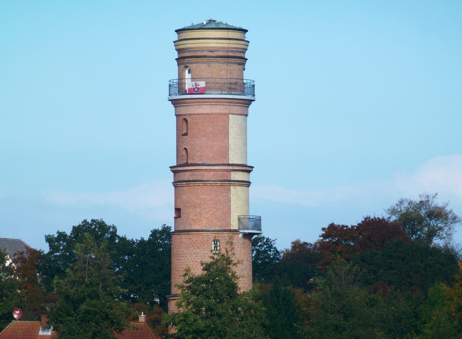 Der Alte Leuchtturm von Travemünde, ein Turm aus rotem Backstein, ragt über herbstlich gefärbte Bäume in den blauen Himmel