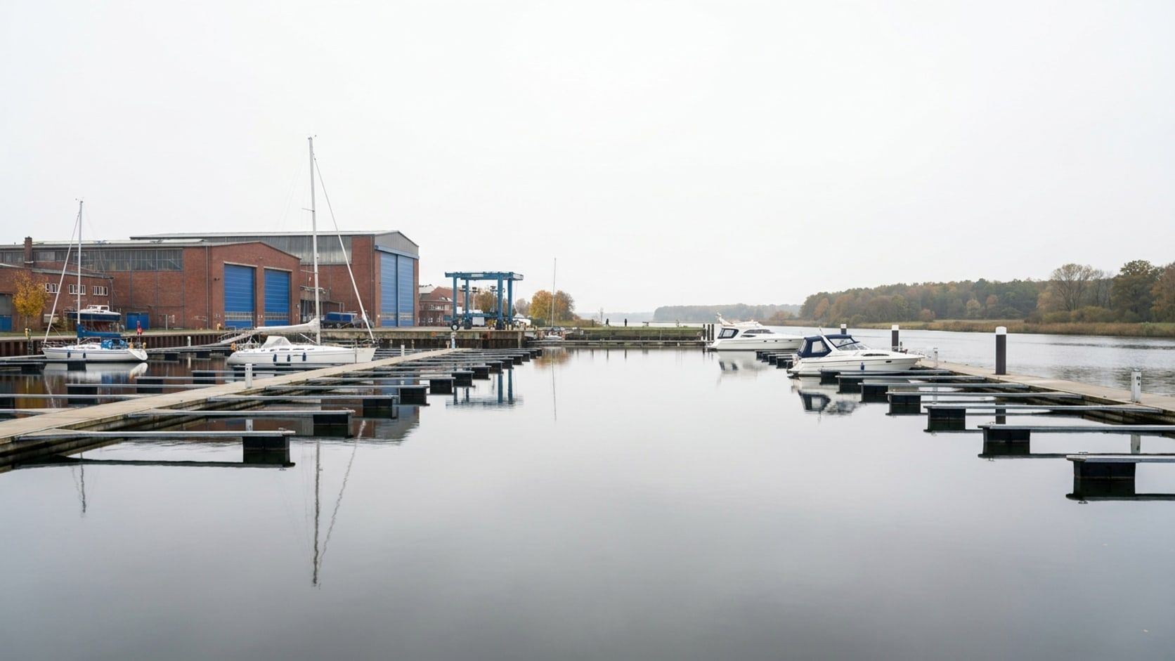 Leere Schwimmstege im Yachthafen an der Trave, wenige Yachten, dahinter rote Werfthallen mit blauen Toren und ein Bootskran, gegenüber bewaldetes Ufer