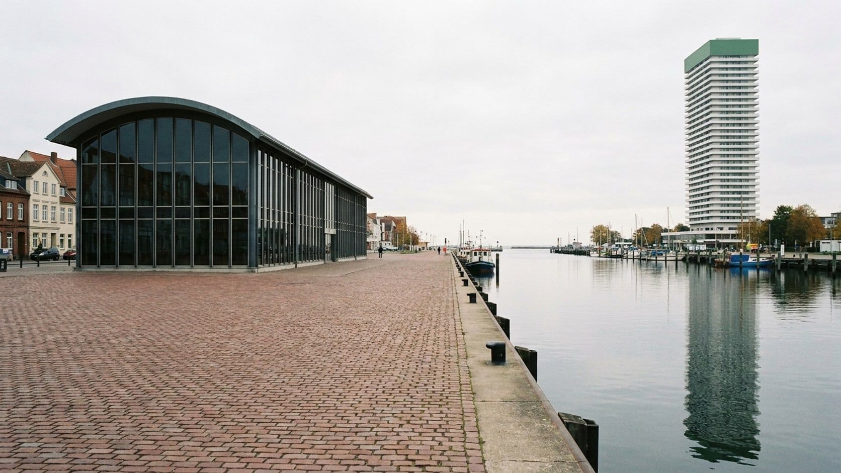 Der leere Ostpreußenkai mit rotbraunem Kopfsteinpflaster, schwarzen Pollern und der gläsernen Halle des Kreuzfahrt-Terminals mit gewölbtem Dach, rechts die Trave, in der Ferne das Hotelhochhaus unter bedecktem Herbsthimmel