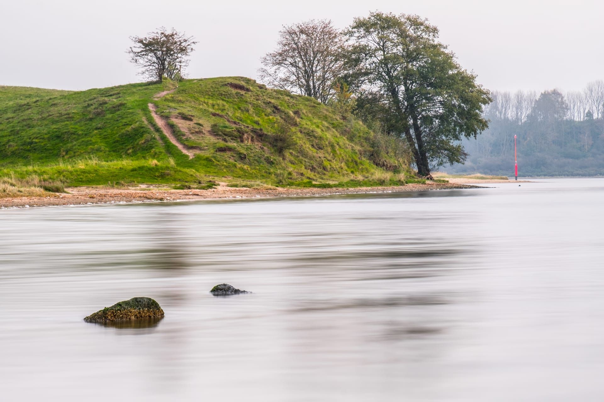 Grüner Hügel mit Bäumen am stillen, milchig glatten Wasser der Trave am Stülper Huk, diesiger Himmel
