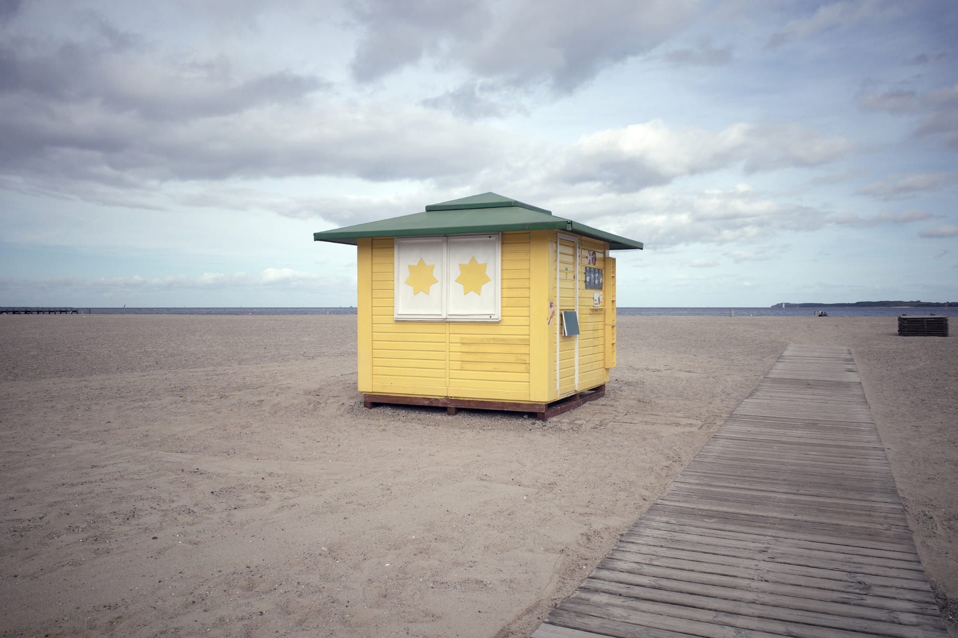 Eine geschlossene gelbe Eisbude am leeren Strand neben einem Bohlenweg, darüber ein wolkiger Himmel