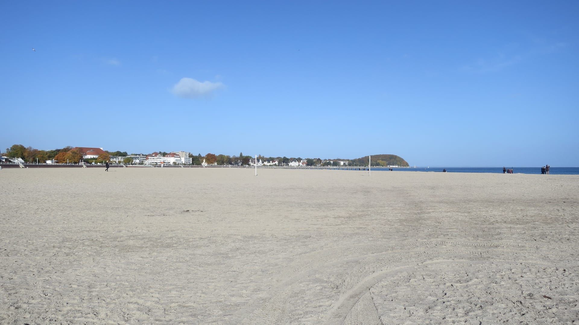 Weiter, fast leerer Sandstrand von Travemünde unter blauem Herbsthimmel, hinten die Strandpromenade und das bewaldete Ufer